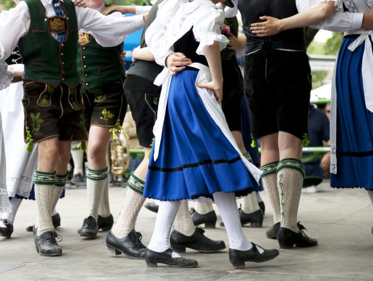A group of people wearing traditional Bavarian clothing dance together outdoors, showing mainly their legs and feet in motion.