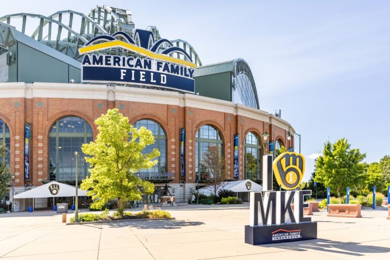 Exterior view of American Family Field stadium with a large “I heart MKE” sign and the Milwaukee Brewers’ logo in the foreground on a sunny day.