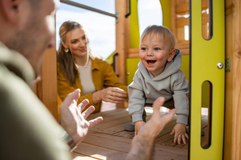 A baby in a gray hoodie crawls toward an adult with open arms, while a woman smiles in the background inside a wooden play structure.