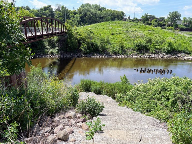 A metal footbridge crosses over a river surrounded by green vegetation; concrete steps lead down to the water’s edge.