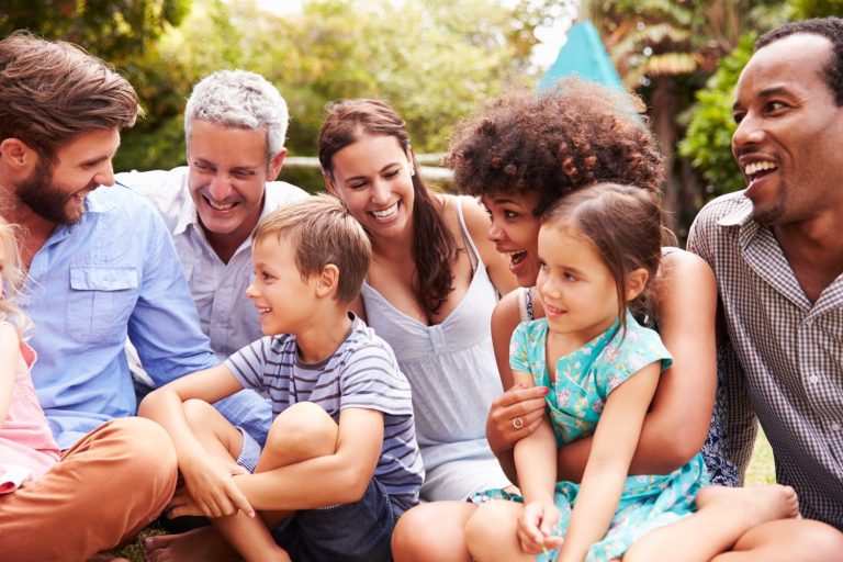 A group of adults and children sit closely together outdoors at Glenwood Manufactured Home Community, smiling and talking on a grassy area with trees in the background.