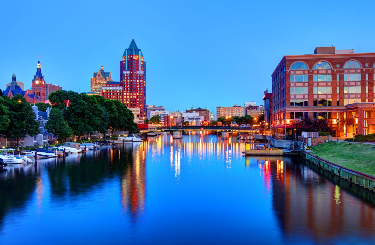 A view of downtown Milwaukee, Wisconsin, at dusk with city buildings illuminated and their reflections visible in the calm river.