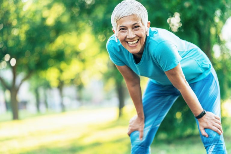 Older adult with short gray hair in athletic wear smiles outdoors, leaning forward with hands on knees in a park setting.