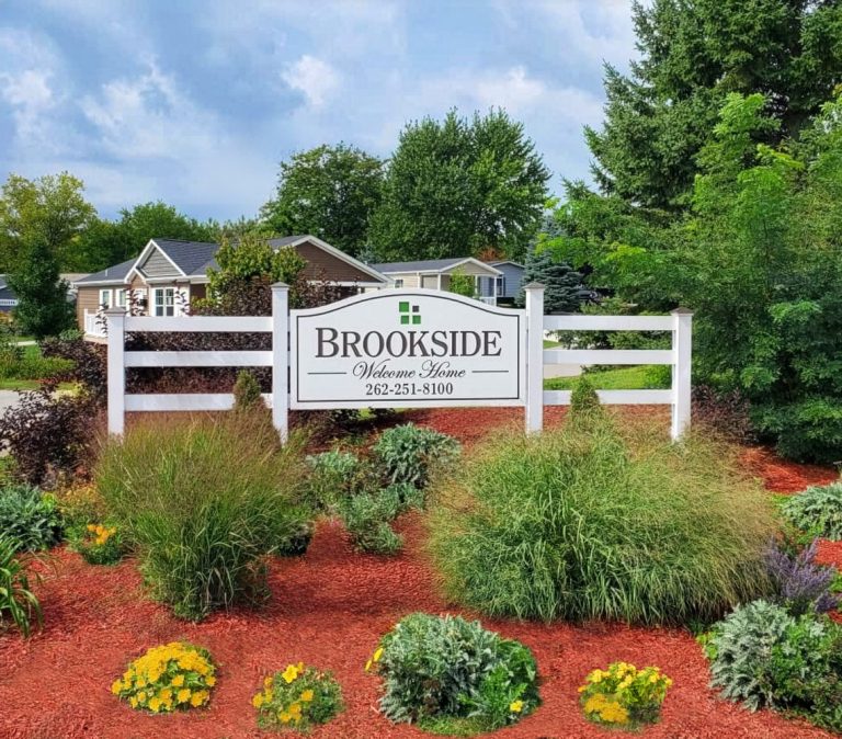 A white sign reading "Brookside Welcome Home" with a phone number, surrounded by landscaped bushes, flowers, and trees, in front of residential houses.