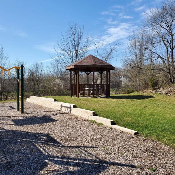 A gazebo stands on a grassy area next to a playground with climbing equipment, under a clear blue sky with leafless trees in the background.